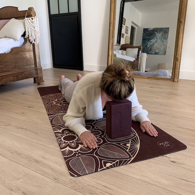 jeune femme allongée sur le ventre au sol sur un tapis de yoga performance bordeaux mandala en posture du corbeau avec la tête posée sur un bloc yoga